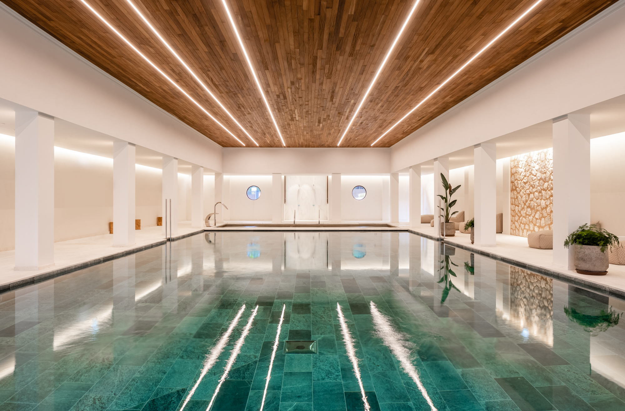An indoor swimming pool with clear turquoise water, surrounded by white walls and columns, featuring wooden ceiling details and ambient lighting
