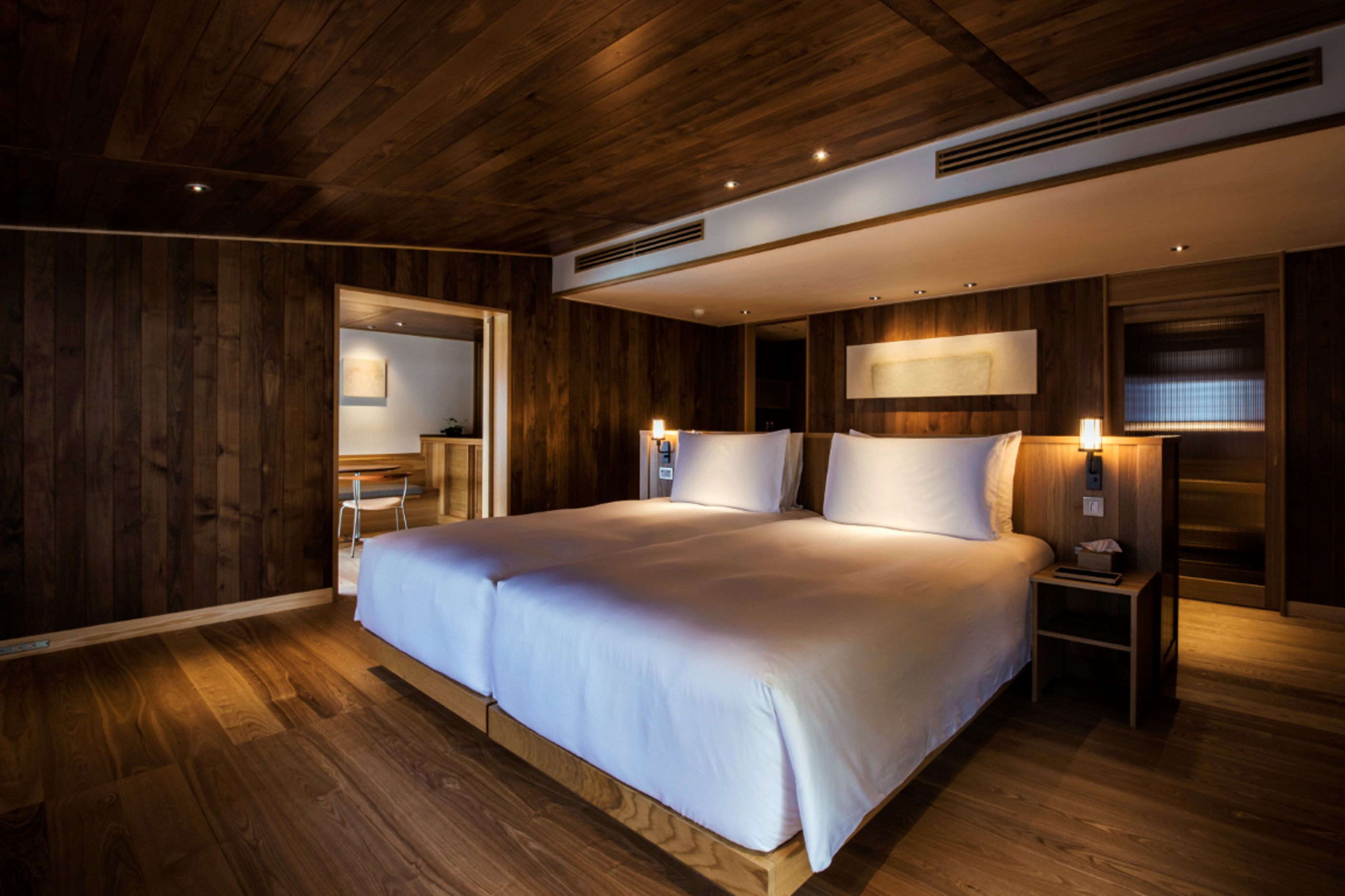 The simplistic bedroom of a suite aboard Guntu featuring a large bed and side lamps with glimpses of a bathroom and seating area