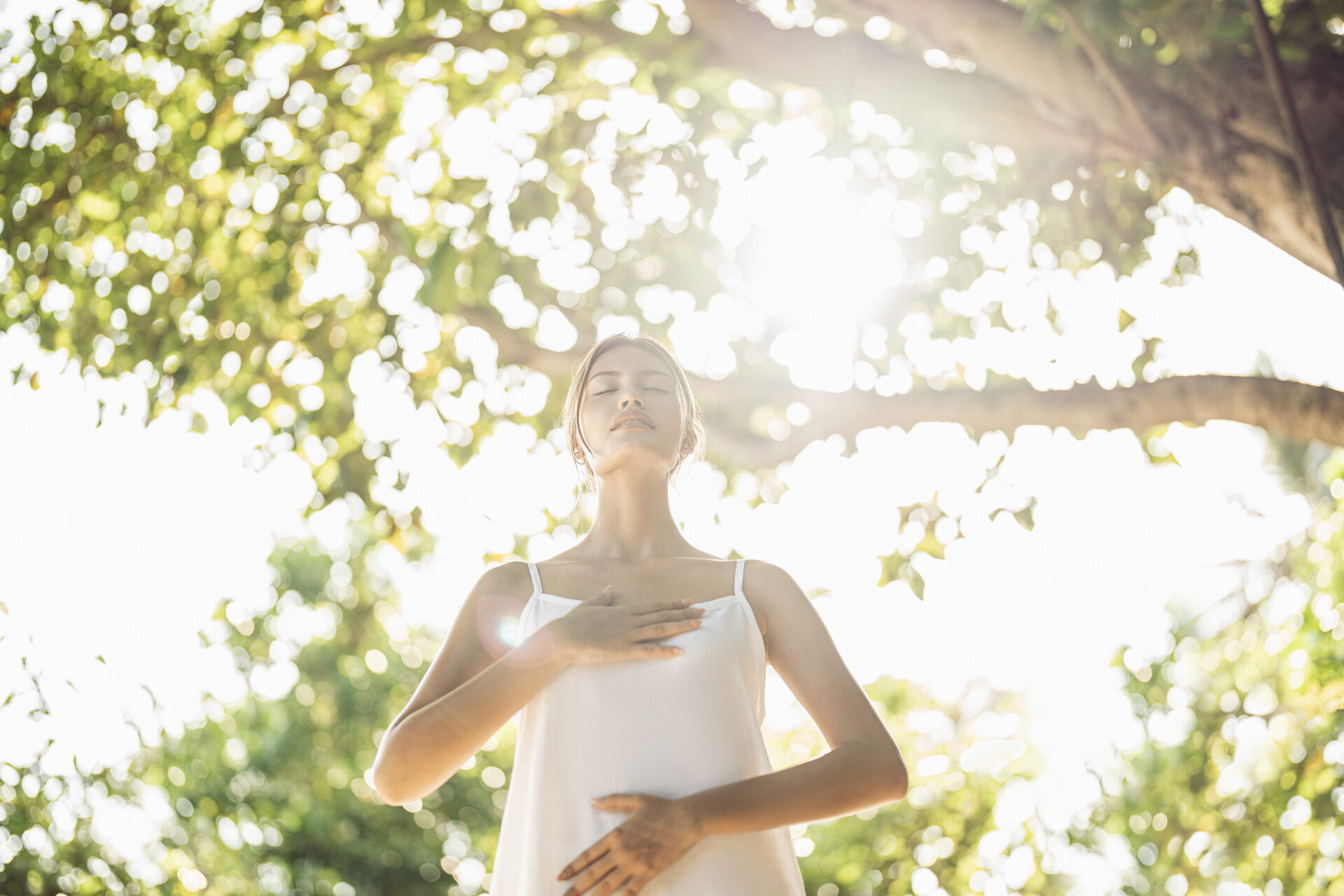 A woman deep breathing with one hand on her heart and one on her stomach beneath a tree