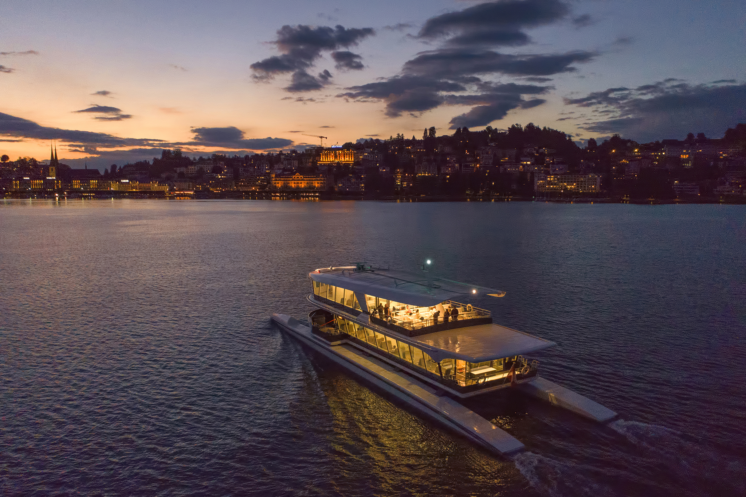 A catamaran sailing along a calm lake at sunset towards an illuminated Lucerne city