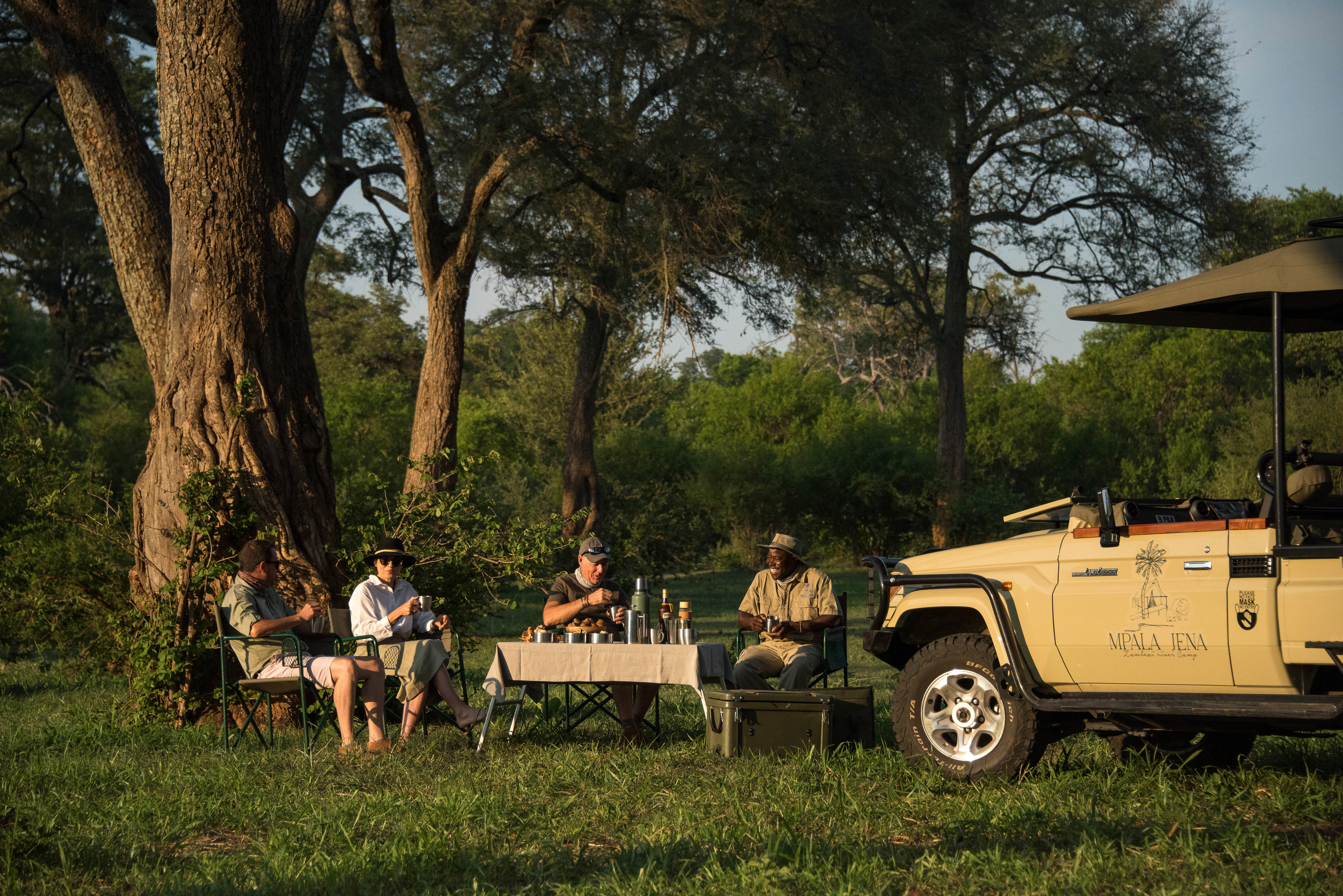 Four people sat beside a safari jeep drinking around a table in the wilderness