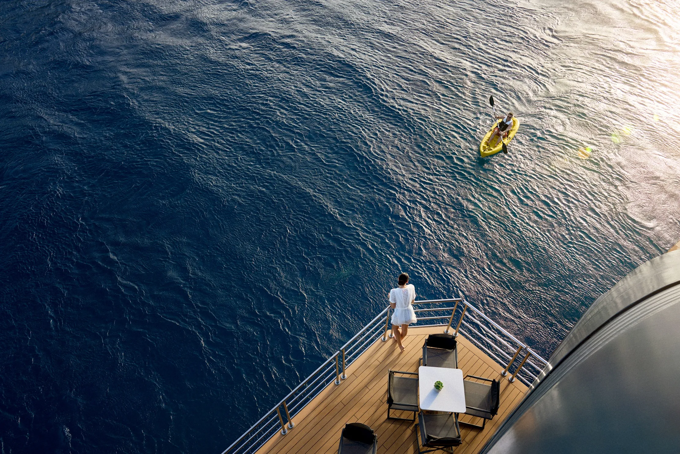 A person stands on a yacht deck overlooking the ocean, with another person kayaking nearby.