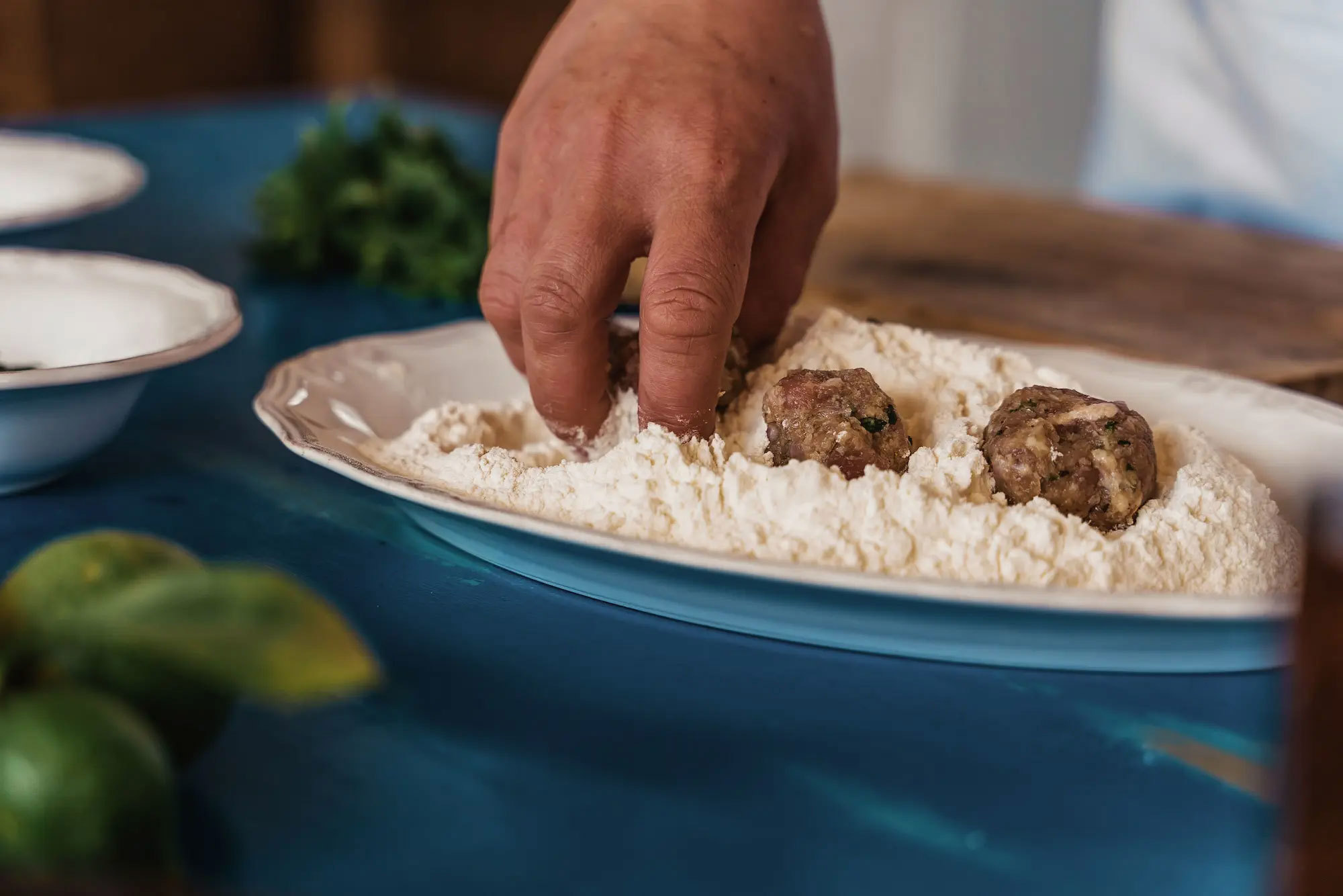 Hand coating meatballs in flour in a blue oblong dish atop a table