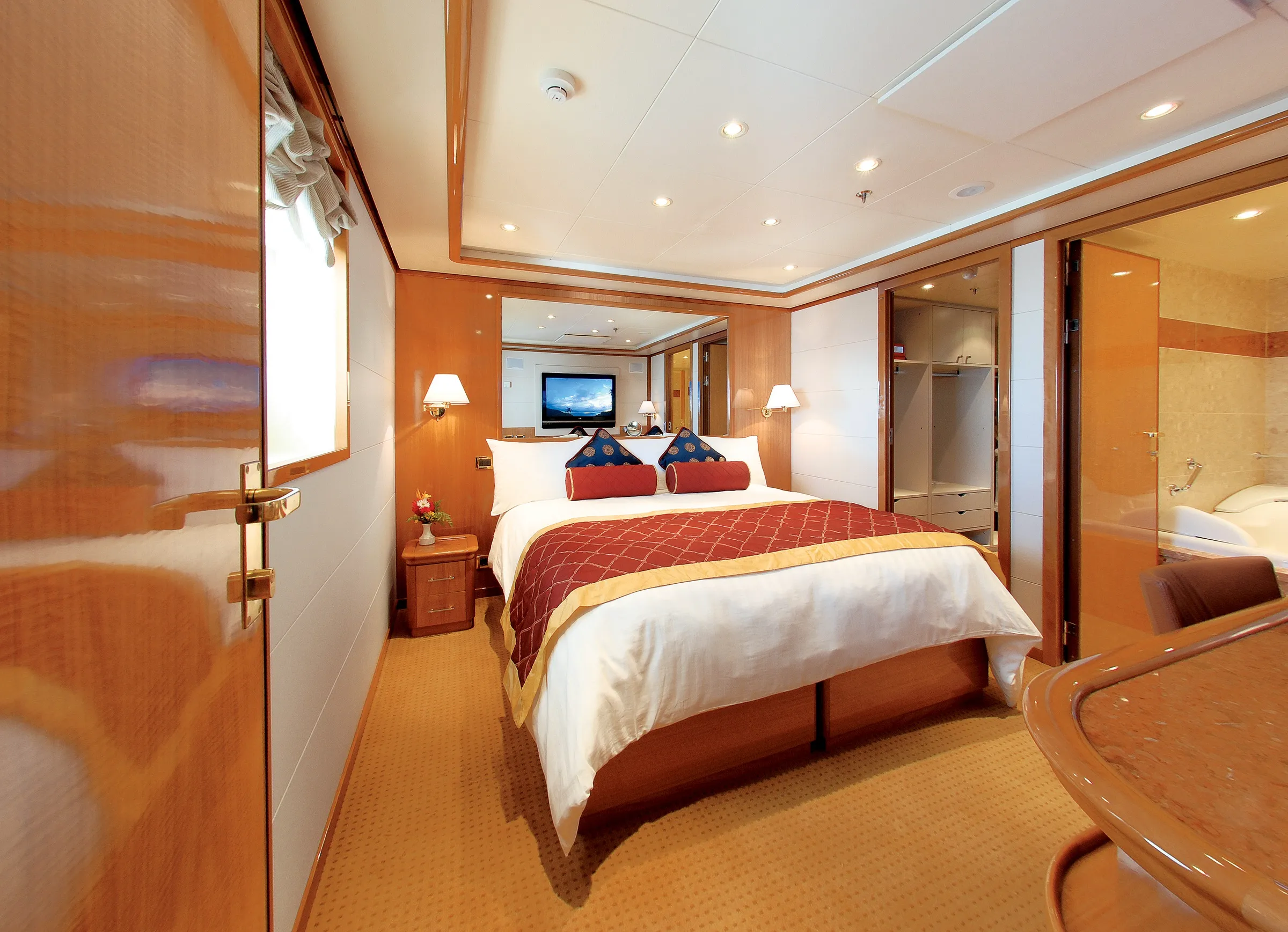 A luxurious hotel room featuring a large bed, nightstands with lamps, a flat-screen TV, and an open door to the bathroom.