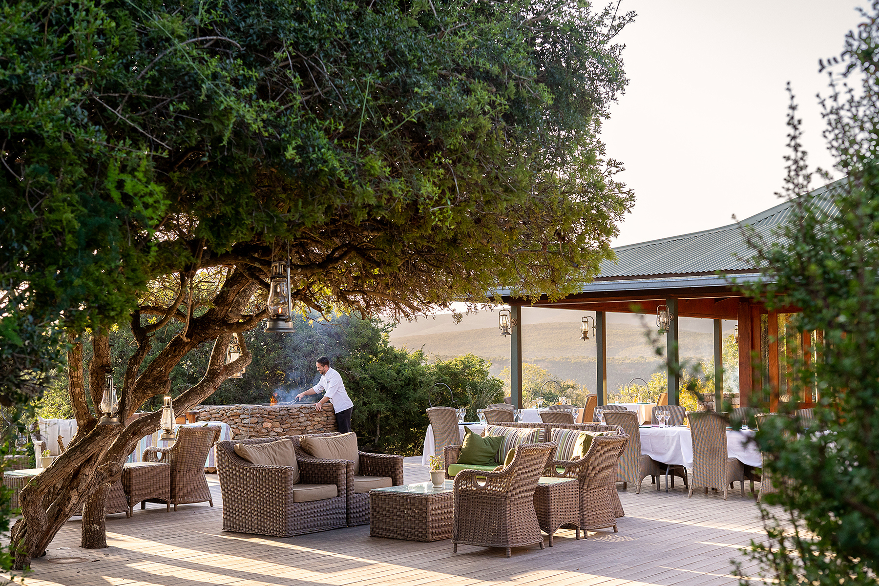 Outdoor seating area of Great Fish River Lodge with rattan furniture shaded by a large tree and chef cooking on the barbecue